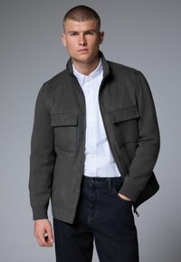 Veste zippée grise en tissu doux avec deux poches poitrine, associée à une chemise blanche et un jean foncé. Texture lisse, design moderne.