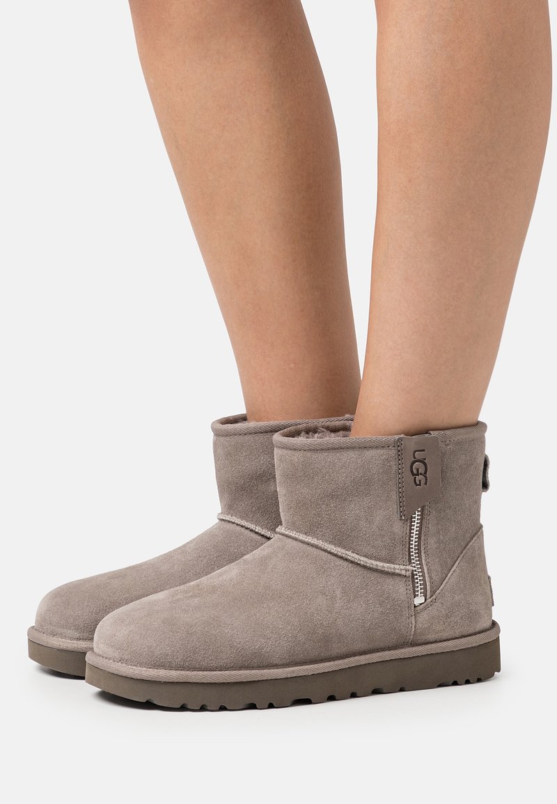 Jambes portant des bottes UGG courtes en daim gris avec des fermetures éclair latérales et des semelles en caoutchouc, présentées sur un fond blanc uni.
