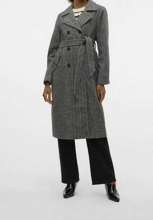 Vrouw draagt een grijze wollen jas in visgraatmotief tot op de knie met dubbele rij knopen, een gebonden riem, zwarte broek en zwarte schoenen met hak.