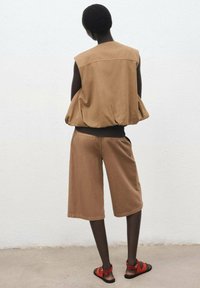 Braunes Baumwoll-Top ohne Ärmel mit gerafftem Saum, kombiniert mit braunen knielangen Shorts und roten Sandalen. Texturierte Oberfläche, lässige Passform.