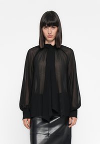 Una donna con capelli scuri lisci indossa una blusa nera trasparente con collo a cravatta e maniche lunghe, abbinata a una gonna di pelle nera, in piedi contro uno sfondo bianco.