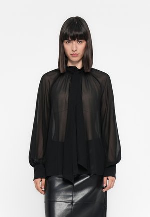 Una donna con capelli scuri lisci indossa una blusa nera trasparente con collo a cravatta e maniche lunghe, abbinata a una gonna di pelle nera, in piedi contro uno sfondo bianco.