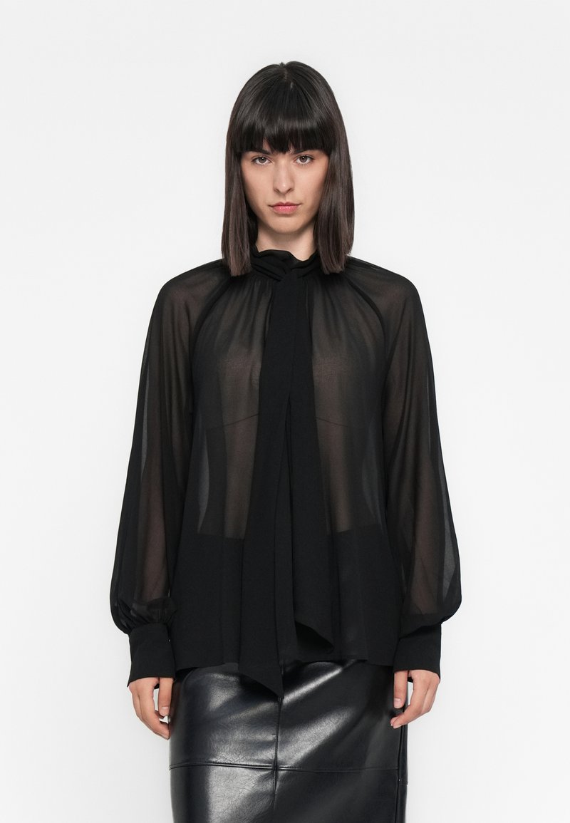 Una donna con capelli scuri lisci indossa una blusa nera trasparente con collo a cravatta e maniche lunghe, abbinata a una gonna di pelle nera, in piedi contro uno sfondo bianco.