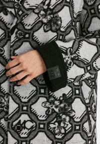 Wollmischung Mantel mit schwarzen Blumen- und geometrischen Mustern, grauem Grundton, schwarzen Manschetten und Knopfverschlüssen. Verfügt über Reißverschlusstaschen.