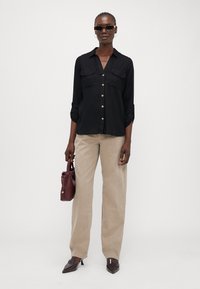 Chemise noire à boutons avec deux poches sur la poitrine, pantalon beige à jambe large, sac à main bordeaux et chaussures foncées à bout pointu. Lunettes de soleil élégantes portées.