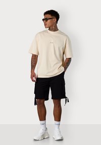 Beige Baumwoll-T-Shirt mit kurzen Ärmeln und kleinem Logo, kombiniert mit schwarzen Cargo-Shorts mit Seitentaschen und Bindebändern sowie weißen Turnschuhen.