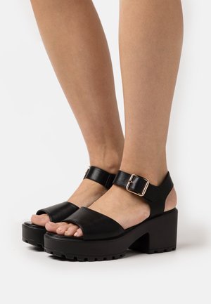 Zwarte leren platform sandalen met een dikke blokhak, een enkelband met een metalen gesp en een open teen ontwerp.