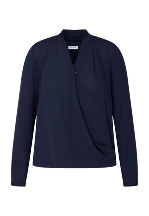 Vrouw die een donkerblauwe blouse met lange mouwen, een V-hals en een gedrapeerd voorpand draagt, gecombineerd met een zwarte broek, handen in de zakken.