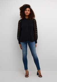 Black blouse with lace sleeves, front buttons, and a structured collar, paired with fitted blue jeans and black heeled sandals.