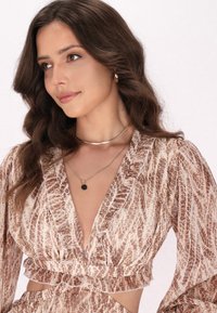 Gelaagde, beige top met bruine krokodillenprint; diepe V-hals, lange pofmouwen, gerimpelde zoom, gelaagd met zilveren ketting en gouden choker.