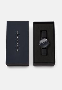 Montre avec bracelet en maille noire, cadran rond, accents argentés et design minimaliste, présentée dans une boîte bleu foncé avec du texte doré.