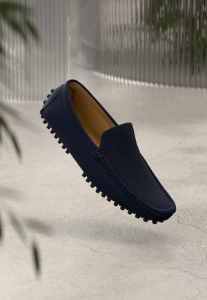 Mocassin à enfiler bleu marine avec semelle cloutée en caoutchouc flottant au-dessus d'un sol en béton clair, avec des plantes floues et un arrière-plan rainuré.