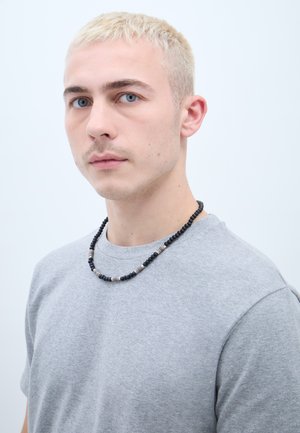 Fekete és ezüst gyöngy nyaklánc, gömbgyöngyökkel, szürke póló felett viselve. A gyöngyözés váltakozó textúrákkal és felületekkel rendelkezik.