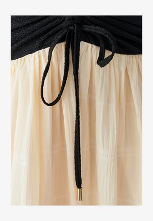 Zwart textuurstof bovenaan met een geplooid ontwerp, overgaand in een zachte, crèmekleurige geplooide rok vastgehouden door een zwarte trekkoord met gouden accent.