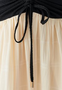 Black textured top with gathered fabric meets a cream, pleated skirt, featuring a black tie with a gold metal tip accent.