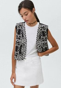 Gilet sans manches noir orné de bijoux et de motifs brodés blancs, associé à un T-shirt blanc uni et une jupe en denim blanche.