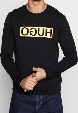 Mężczyzna w czarnej koszuli z długim rękawem z żółtym prostokątnym logo z odwróconym napisem "HUGO", ubrany w ciemnoniebieskie dżinsy, ręce złożone z przodu.