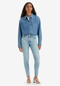 Ljusblå denimskjorta med långa ärmar, två frontfickor och knappstängning, tillsammans med ljusblå skinny jeans och vita blockklackssandaler.