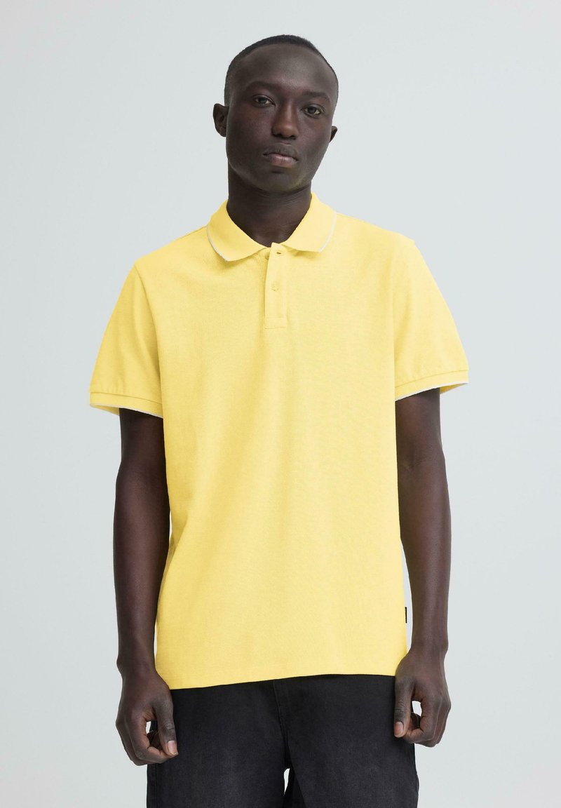 Homme à la peau foncée portant un polo à manches courtes jaune et un pantalon noir, debout devant un fond gris clair.