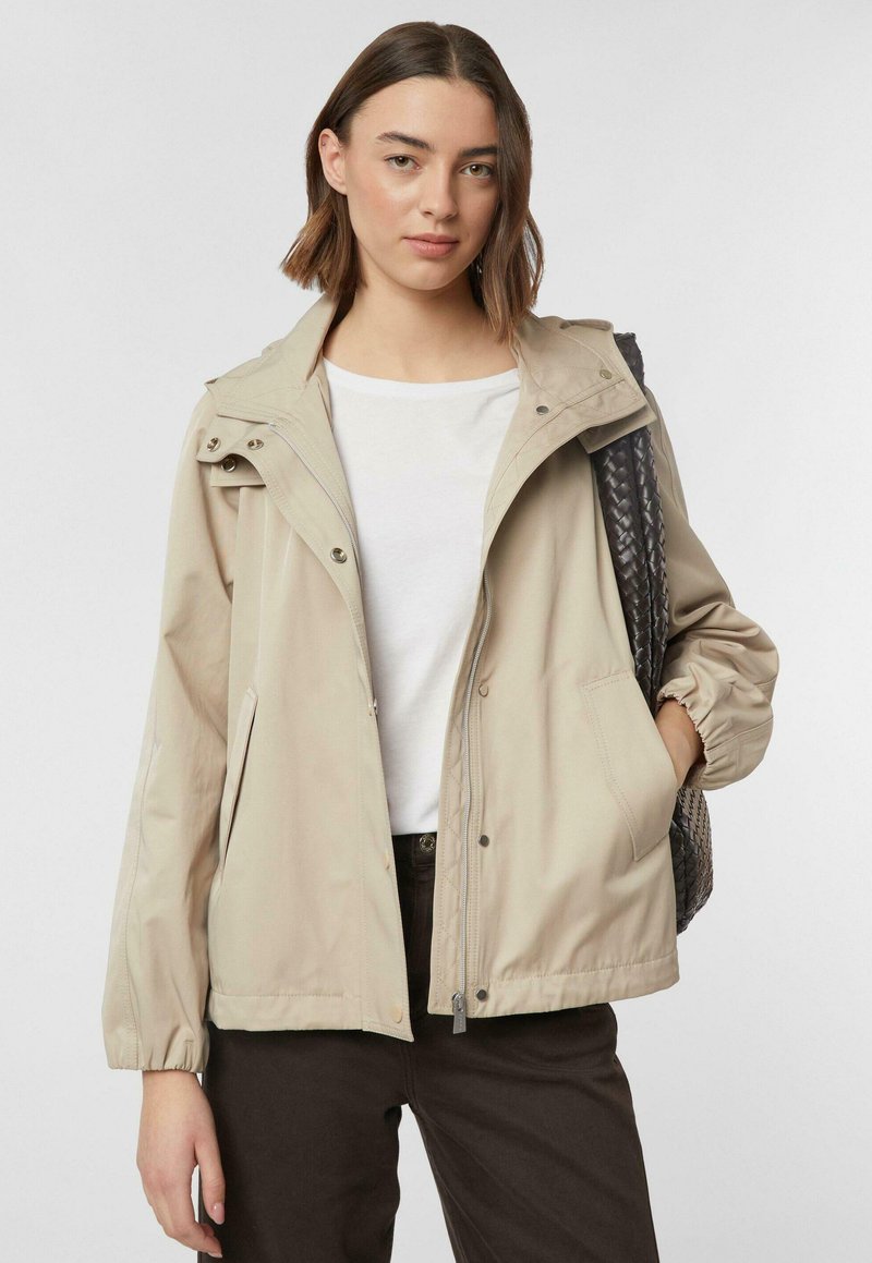 Junge Frau trägt beige Jacke, weißes Hemd, dunkle Hose, trägt eine strukturierte schwarze Tasche über der Schulter und posiert vor neutralem Hintergrund.