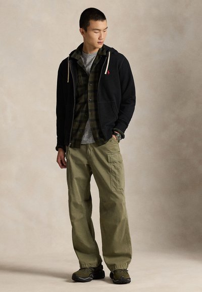 Moški stoji, oblečen v črno jopa z zadrgo, zeleno karirano srajco čez sivo majico, olivno zelene hlače cargo in temne superge na enobarvnem bež ozadju.