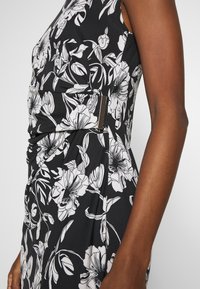 Sukienka w kwiaty z czarnym tłem i białymi wzorami, z marszczeniami oraz poziomym metalicznym akcentem z boku.