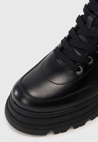 Primer plano de una bota de cuero negro que muestra una suela gruesa y texturizada, puntera redondeada, detalles cosidos y cordones negros trenzados a través de ojetes de metal.