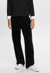 Mustat corduroy-housut suoralla lahkeella, joissa on pystyribbaus ja etutaskut, yhdistettynä valkoisiin lenkkareihin ja mustaan paitaan.