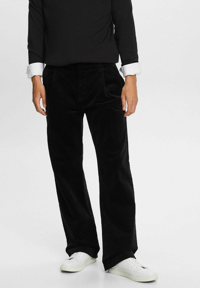 Mustat corduroy-housut suoralla lahkeella, joissa on pystyribbaus ja etutaskut, yhdistettynä valkoisiin lenkkareihin ja mustaan paitaan.