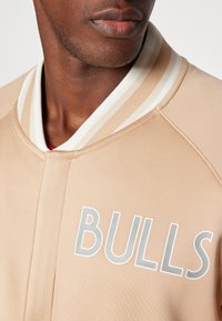 Bež sportski sako s rebrastim bijelim i smeškim kragnom. Ima veliki sivi tekst "BULLS" napred. Glatka tekstura tkanine.