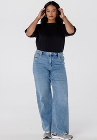 Jonge vrouw die haar aanpast, gekleed in een zwart top, wijde blauwe jeans, gouden ring oorbellen en grijze sneakers, staande tegen een effen witte achtergrond.