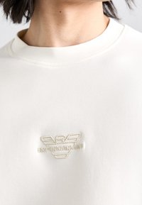 Sudadera de algodón blanca con cuello redondo que presenta un logotipo bordado de Emporio Armani en beige en el pecho. Textura suave, diseño minimalista.