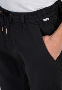 Pantalones negros de cintura elástica con cordón; bolsillos laterales; la tela parece suave con una ligera textura; incluye una pequeña etiqueta de la marca.