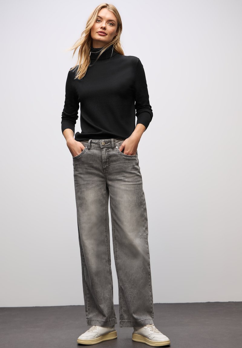 Maglione nero a collo alto con maniche lunghe abbinato a jeans larghi grigi chiari. Sneakers bianche completano l'outfit, caratterizzate da una texture liscia.