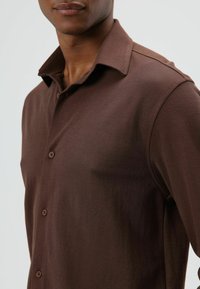 Man draagt een bruin overhemd met knopen en een kraag, weergegeven van nek tot midden van de torso tegen een effen achtergrond.