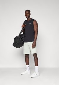 Camiseta sin mangas negra, pantalones cortos verde claro, zapatillas blancas y bolsa de deporte negra; diseño limpio, colores contrastantes y logo visible en la ropa.