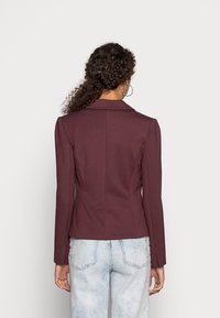 Blazer ajusté bordeaux à manches longues et col classique, présentant une texture lisse et une coupe sur mesure. Porté avec un jean en denim clair.