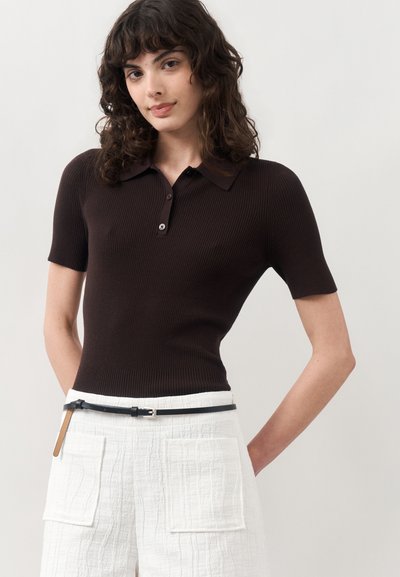 Femme aux cheveux bouclés et foncés portant un polo côtelé marron foncé ajusté et un pantalon blanc texturé avec des poches plaquées devant et une fine ceinture noire.