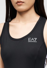 Κοντινό πλάνο γυναίκας που φοράει μαύρο αμάνικο αθλητικό τοπ με λευκό λογότυπο EA7 Emporio Armani στο στήθος.