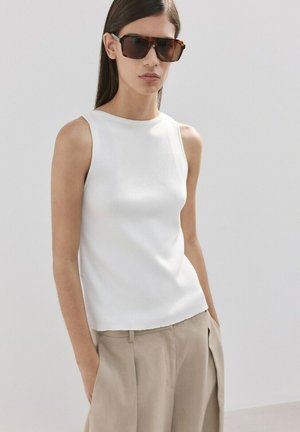 Femeie purtând un tricou alb fără mâneci, pantaloni bej cu talie înaltă și mâinile în buzunare, cu ochelari de soare mari maro, pe un fundal simplu.