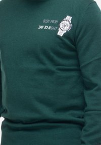 Maglione lavorato a maglia verde scuro con grafica di un orologio stampata e il testo "IMPEGNATO DAL GIORNO ALLA NOTTE." Polsini e orlo a coste con texture liscia.