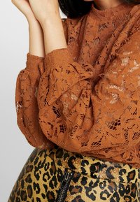 Frau trägt eine rostfarbene, florale Spitzenbluse mit hohem Kragen und Ballonärmeln, kombiniert mit einem Leopardenmuster-Rock mit Vorderzipper.