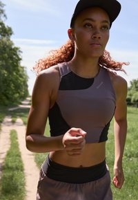 Sportlicher Crop-Top aus dunkelgrauem und schwarzem Stoff, kombiniert mit elastischen Shorts. Läufer in Bewegung auf einem Schotterweg, umgeben von Grün.