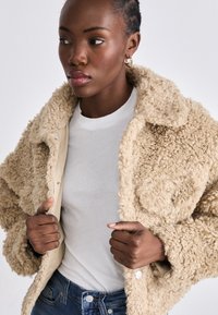 Beige faux fur jakke med krage, knappelukking og to frontlommer. Brukt over en hvit t-skjorte med teksturert stoff.