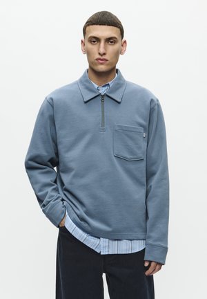 Blauwe rits-hoodie met een platte kraag, borstzak en geribbelde manchetten, gedragen over een lichtblauw gestreept overhemd met knopen.