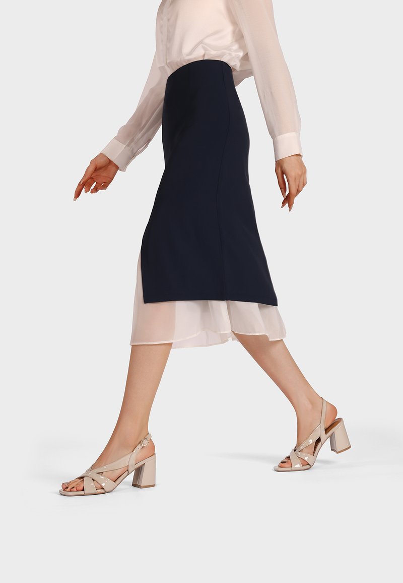Donna che cammina indossando una gonna midi blu navy sovrapposta a un abito bianco trasparente con sandali beige con tacco largo, su uno sfondo semplice.