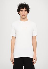 Hvid t-shirt med korte ærmer lavet af bomuld, med et diskret EA7-logo i guld på brystet. Enkel rund halsudskæring og afslappet pasform.