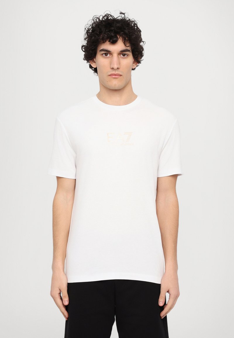 Hvid t-shirt med korte ærmer lavet af bomuld, med et diskret EA7-logo i guld på brystet. Enkel rund halsudskæring og afslappet pasform.