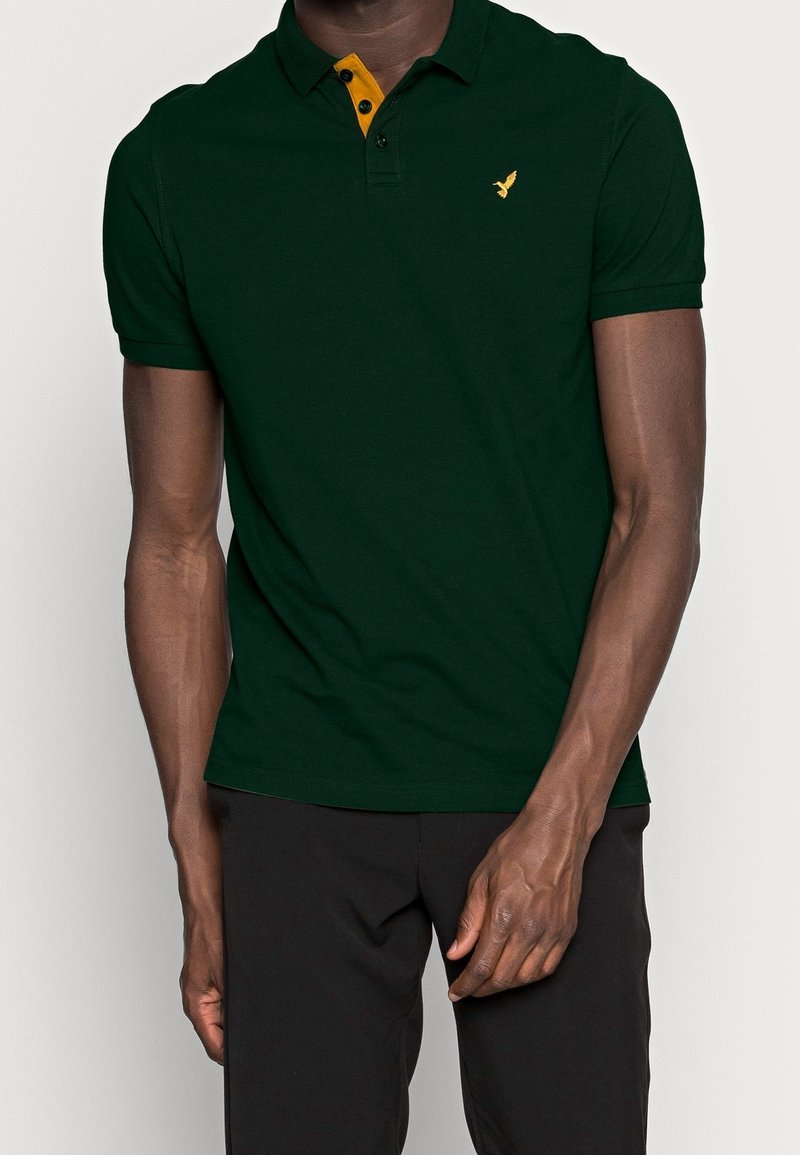 Homme portant un polo vert foncé avec col intérieur jaune et logo d'oiseau sur la poitrine, associé à un pantalon noir sur un fond uni.