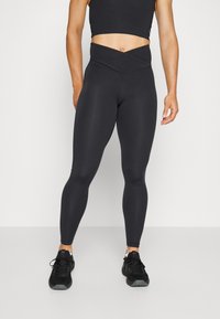 Black high-waisted leggings with a crossover waistband design, made of smooth, stretchy fabric, paired with black athletic shoes.
